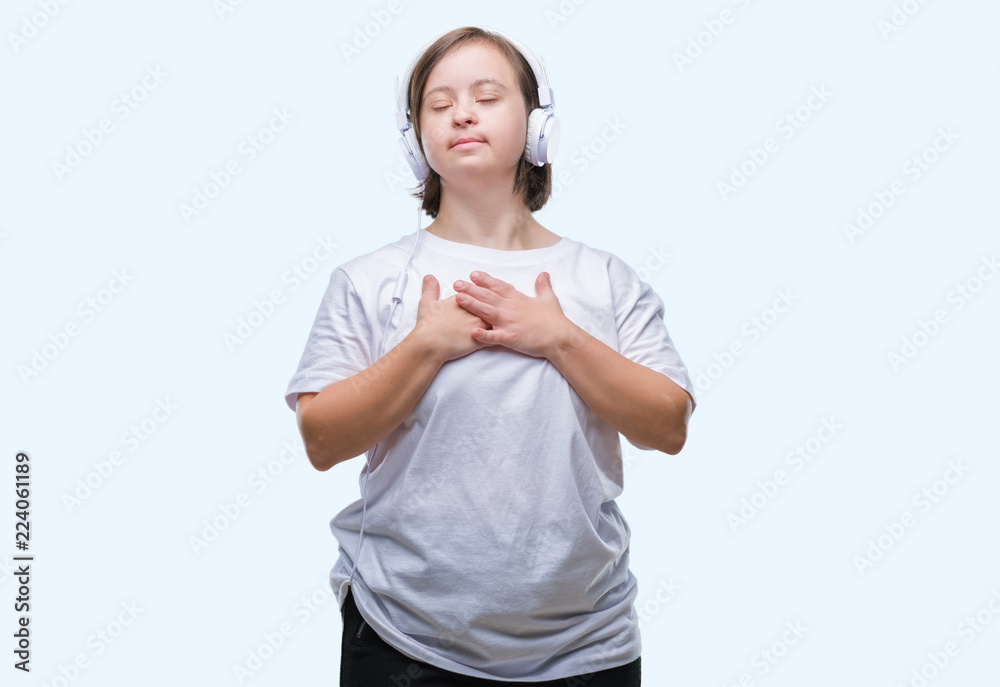 Young adult woman with down syndrome wearing headphones over isolated background smiling with hands on chest with closed eyes and grateful gesture on face. Health concept. Young adult woman with down syndrome wearing headphones over isolated background smiling with hands on chest with closed eyes and grateful gesture on face. Health concept.