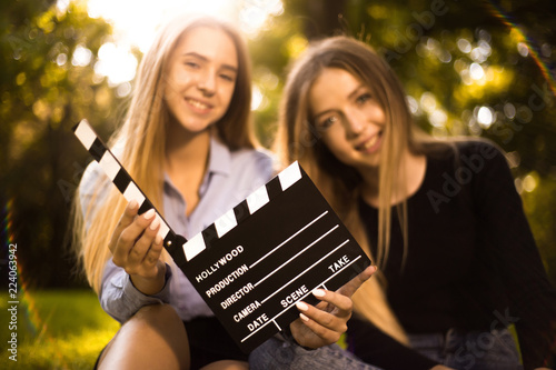 Happy girls sisters students sitting in the park outdoors on grass have a rest holding film making clapperboard. Focus on clapperboard.