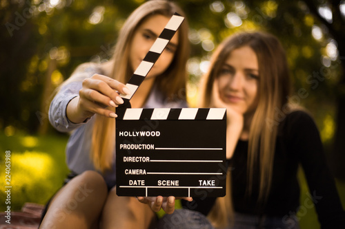 Happy girls sisters students sitting in the park outdoors on grass have a rest holding film making clapperboard. Focus on clapperboard.