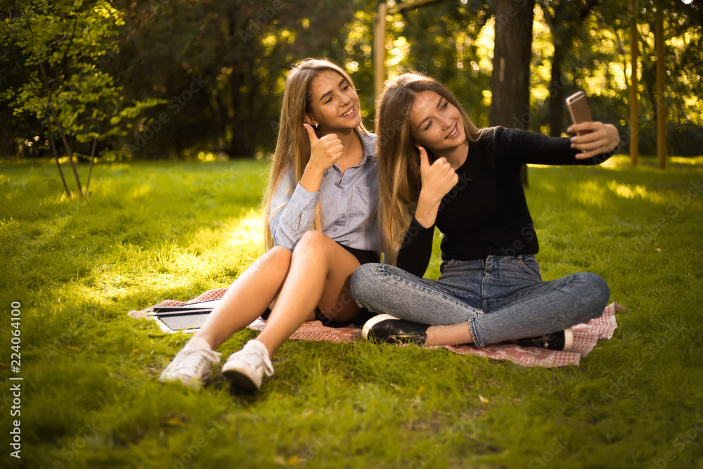 Happy girls sisters students sitting in the park outdoors on grass have a rest take a selfie by mobile phone showing thumbs up gesture. Happy girls sisters students sitting in the park outdoors on grass have a rest take a selfie by mobile phone showing thumbs up gesture.