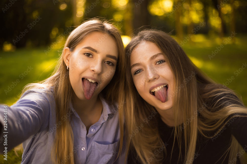 Happy laughing girls sisters students sitting in the park outdoors on grass have a rest take a selfie by camera showing tongue. Happy laughing girls sisters students sitting in the park outdoors on grass have a rest take a selfie by camera showing tongue.