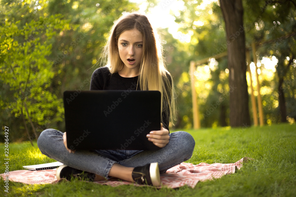 Displeased girl student sitting in the park outdoors on grass using laptop computer typing with mouth opened. Displeased girl student sitting in the park outdoors on grass using laptop computer typing with mouth opened.