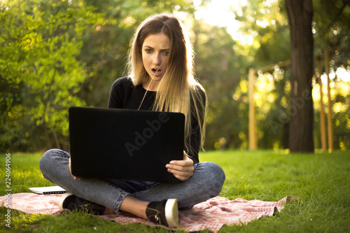 Displeased girl student sitting in the park outdoors on grass using laptop computer typing with mouth opened.