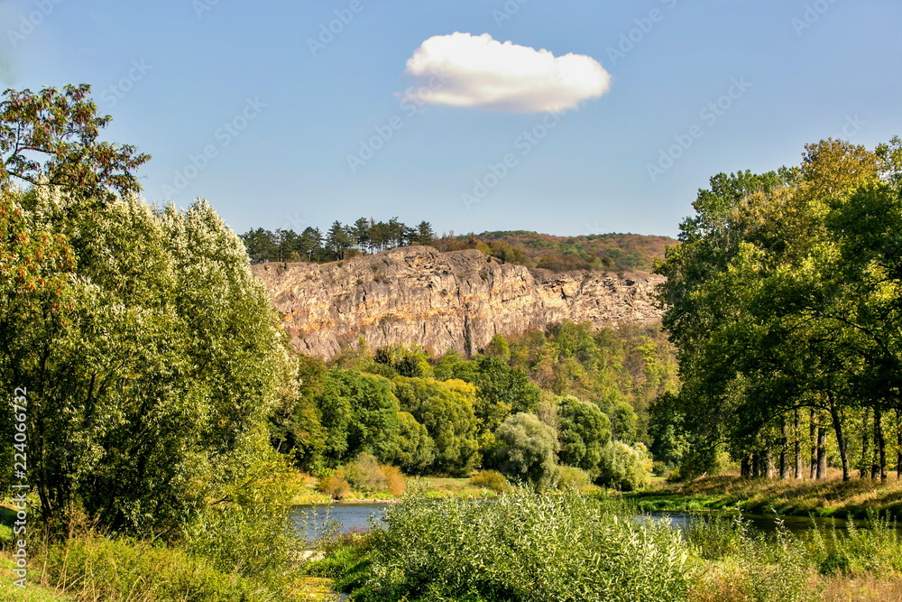 View of valley with river and high brown pink steep limestone rock, trees lined on the top of the rock, white cloud in the middle of blue sky, bright fall sunny day, bushes in foreground