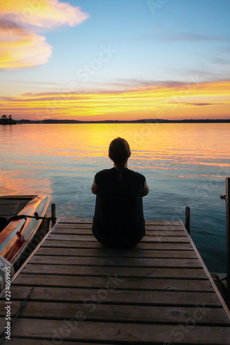 Beautiful sunrise sunset scene of a young woman sitting at the end of a pier looking at the colorful sky. Concepts of vacation, travel, tranquility