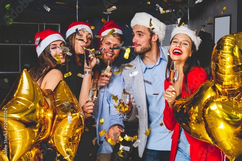 Celebrating New Year together. Group of beautiful young people in Santa hats throwing colorful confetti and looking happy.