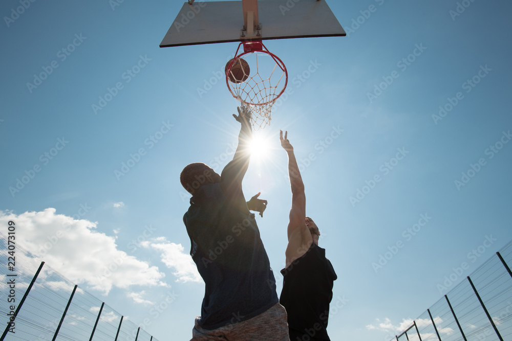Naklejka premium Low angle view of two young men playing basketball and jumping by hoop against blue sky, copy space