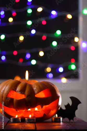 Halloween pumpkin. Candles, bat and glowing pumpkin. Harvest for Halloween with colorful bokeh. Atmosphere for holiday. Pumpkin on wooden boards against background of colored garlands