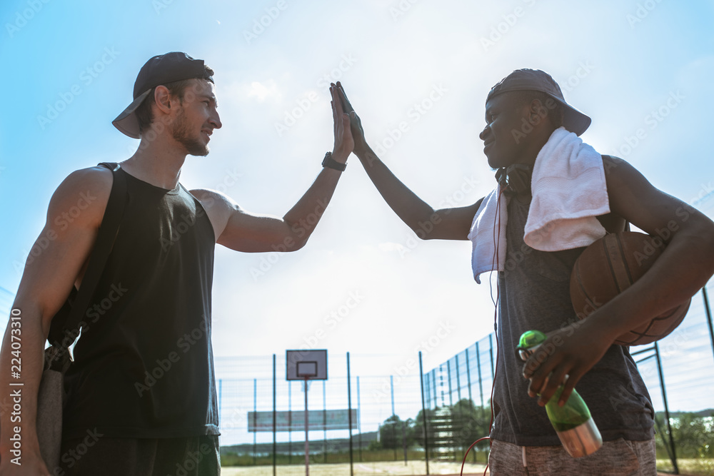 Fototapeta premium Side view portrait of two basketball players doing high-five against blue sky after match in outdoor court, copy space