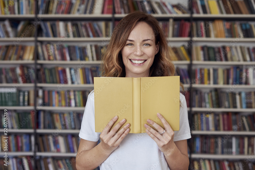 Portrait of smiling woman model with opened book in a library, bookshelf behind, long hair. Hipster college student lady.. Portrait of smiling woman model with opened book in a library, bookshelf behind, long hair. Hipster college student lady..