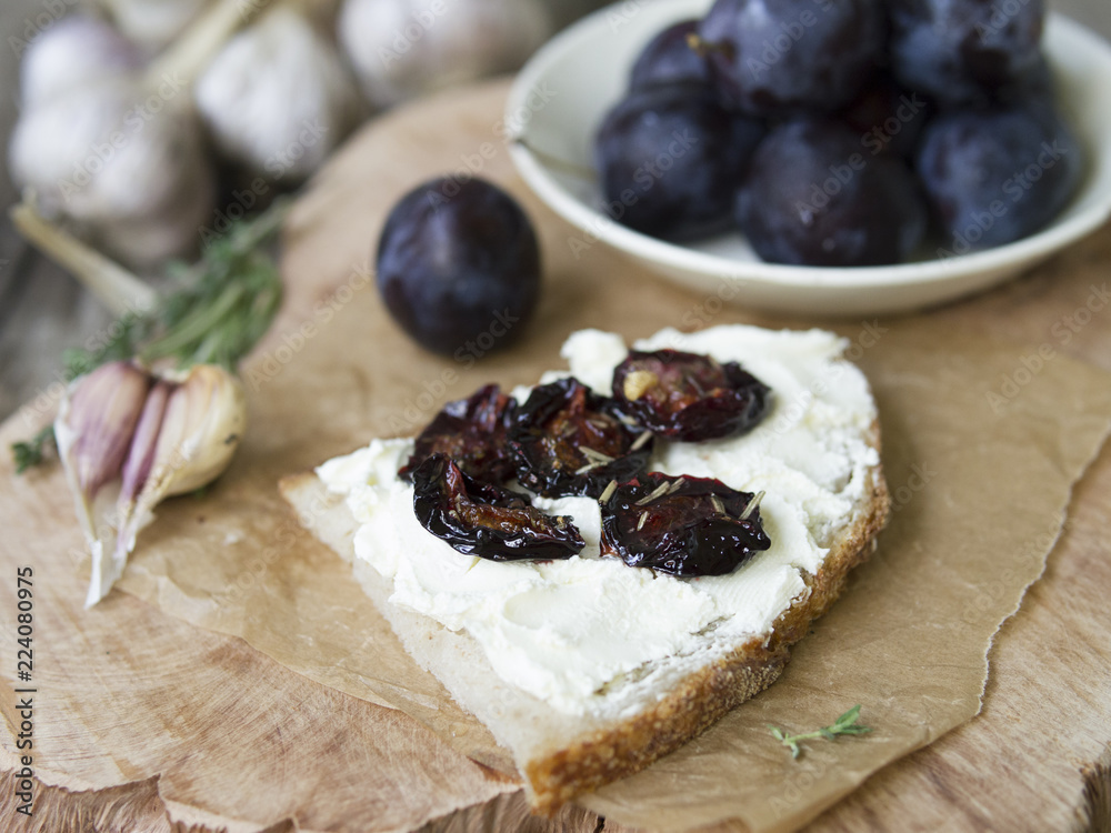 Glass jar with dried plums and fresh rosemary on wooden serving board, selective focus. Sandwich with gray bread, cream cheese and dried plums on a wooden board. Copy space, close up