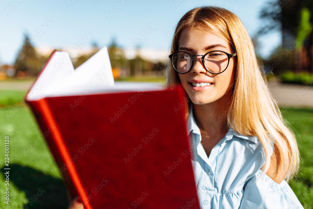 Portrait of a cheerful beautiful student girl sitting outdoors on green grass, wearing glasses, holding a book and reading it Portrait of a cheerful beautiful student girl sitting outdoors on green grass, wearing glasses, holding a book and reading it
