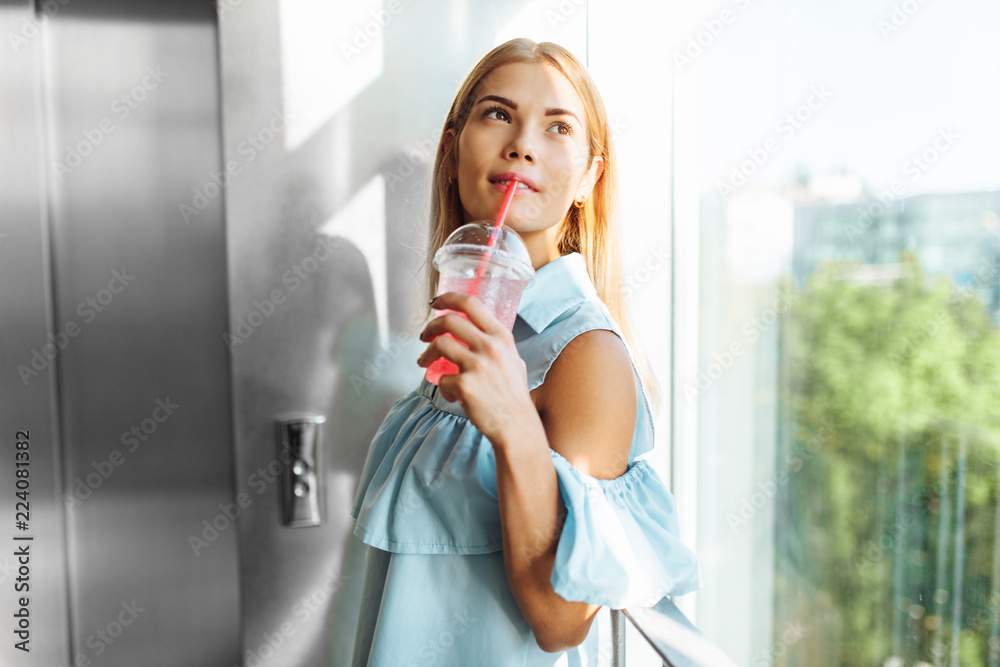 Portrait of a beautiful girl standing near the window in the room, with a cocktail in his hands Portrait of a beautiful girl standing near the window in the room, with a cocktail in his hands