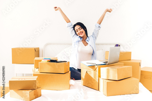 Successful young woman freelancer with arms up working and use laptop computer with cardboard box on bed at home - SME business online and delivery concept