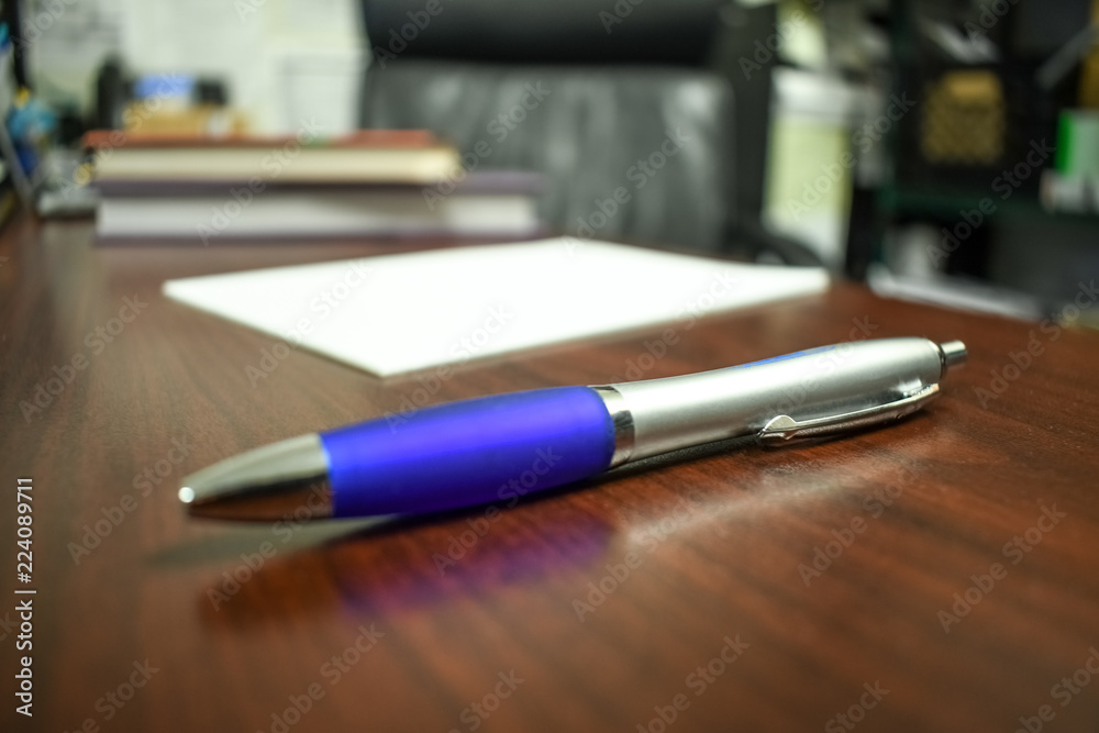 A pen sits in the focus surrounded by the office it never leaves on a dark brown desk next to a black leather chair right in front of some textbooks. A white note pad waiting to be written on A pen sits in the focus surrounded by the office it never leaves on a dark brown desk next to a black leather chair right in front of some textbooks. A white note pad waiting to be written on