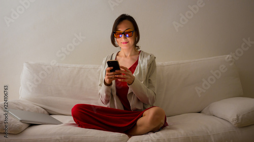 Young woman is sitting on white sofa looking into the smartphone with yellow (amber) blue light blocking eye glasses - health and wellness concept for melatonin and circadian rhythm