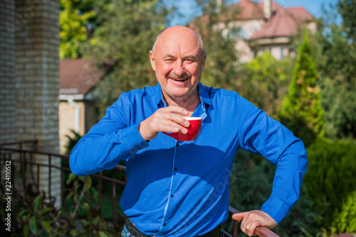 Fototapeta Naklejka Na Ścianę i Meble -  Senior man at morning. Holding a cup of tea in hands.  Man wearing a blue shirt posing at the camera at home yard. Old age, drink and people concept - close up of happy senior man with cup of tea  