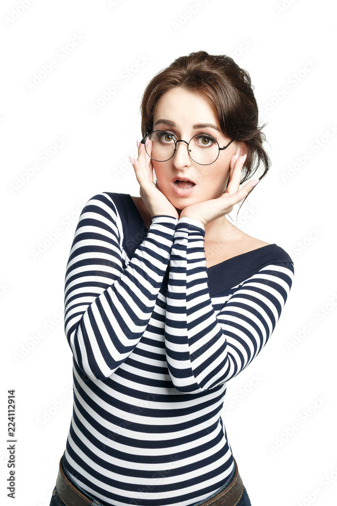 A young woman in a striped tight sweater, wearing round glasses, makes a regrettable face, holding her hands to her face. Isolated on white background. wide open eyes and mouth. A young woman in a striped tight sweater, wearing round glasses, makes a regrettable face, holding her hands to her face. Isolated on white background. wide open eyes and mouth.