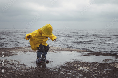  A boy in a yellow raincoat jumps on puddles on the beach.