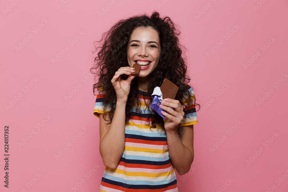 Photo of lovely woman 20s with curly hair eating chocolate bar, isolated over pink background Photo of lovely woman 20s with curly hair eating chocolate bar, isolated over pink background