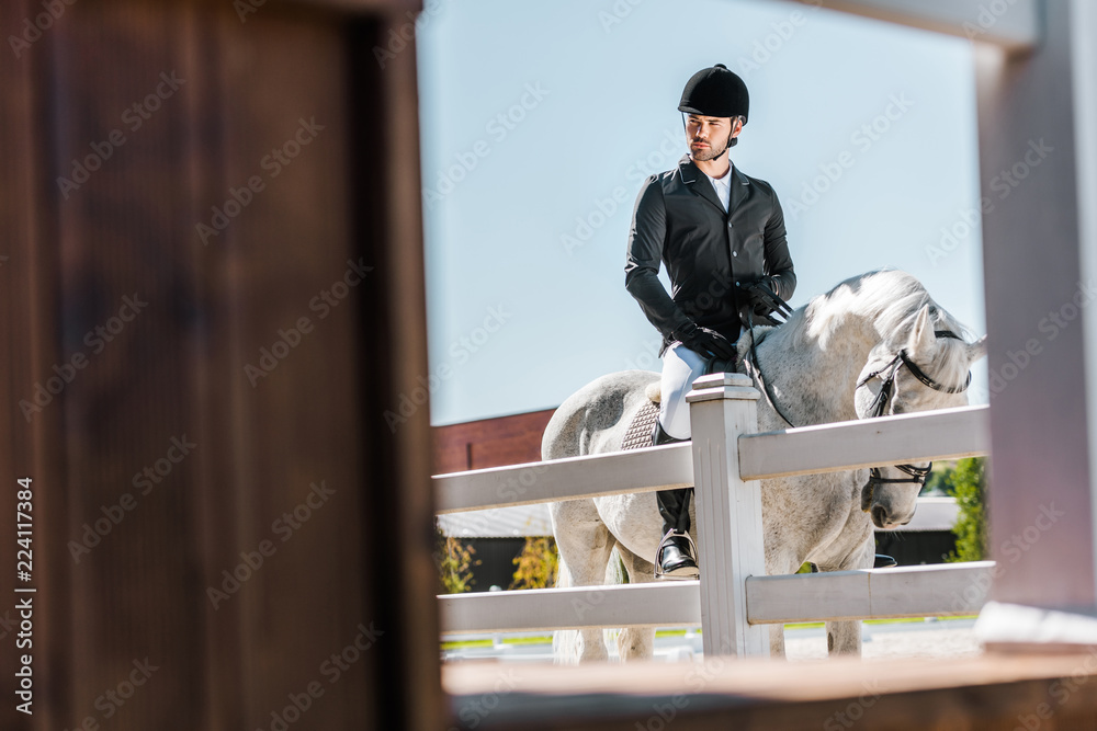view through fence on handsome male equestrian in professional apparel sitting on horse at ranch