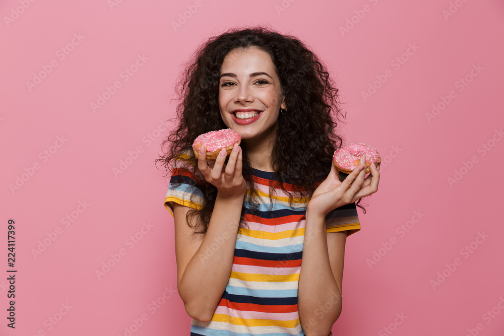 Photo of european woman 20s with curly hair playing around and eating donuts, isolated over pink background