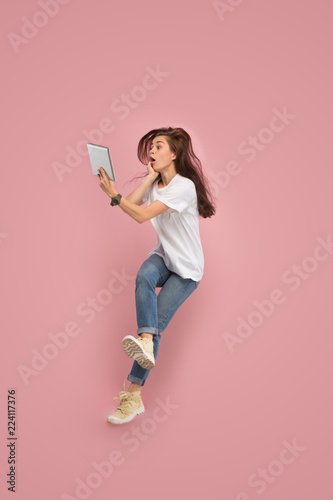 Gadget in modern life. Jump of young woman over pink studio background using laptop or tablet gadget while jumping. Runnin surprised girl in motion or movement. Human emotions and facial expressions
