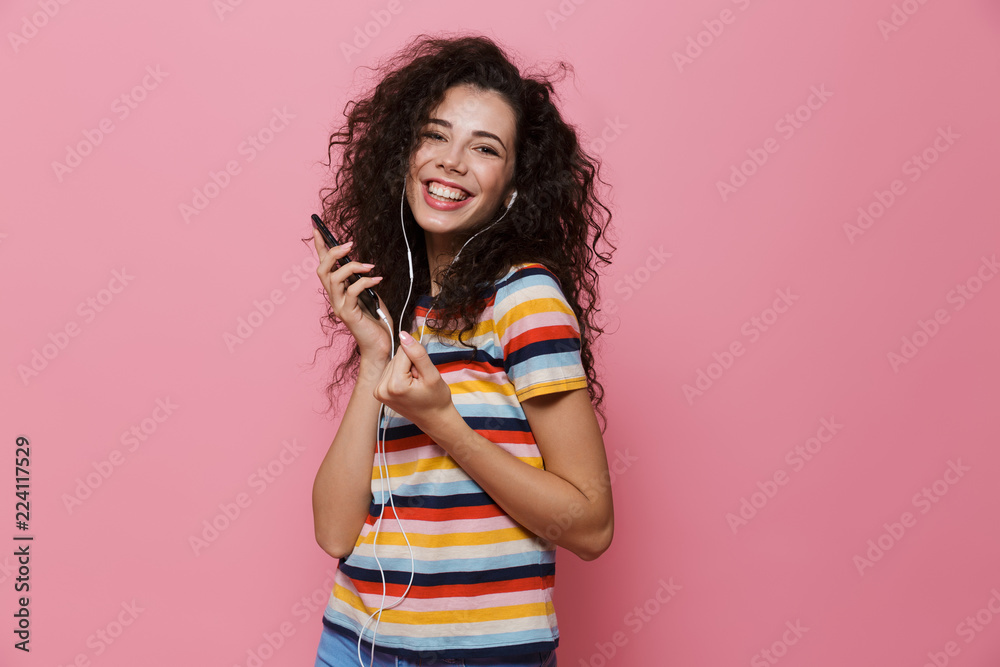 Image of smiling woman 20s with curly hair holding smartphone and listening to music via headphones, isolated over pink background Image of smiling woman 20s with curly hair holding smartphone and listening to music via headphones, isolated over pink background