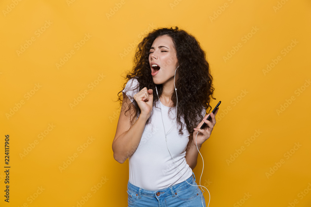 Obraz premium Image of happy woman 20s with curly hair singing while holding smartphone and listening to music via headphones, isolated over yellow background