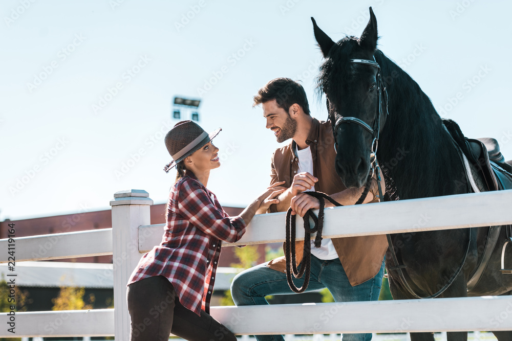 smiling female and male equestrians standing near fence with horse and looking at each other at ranch - obrazy, fototapety, plakaty 