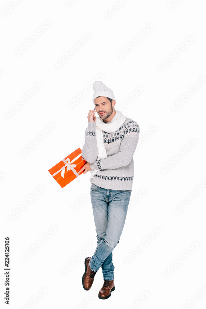 smiling young man in sweater, scarf and hat holding gift box and talking by smartphone isolated on white