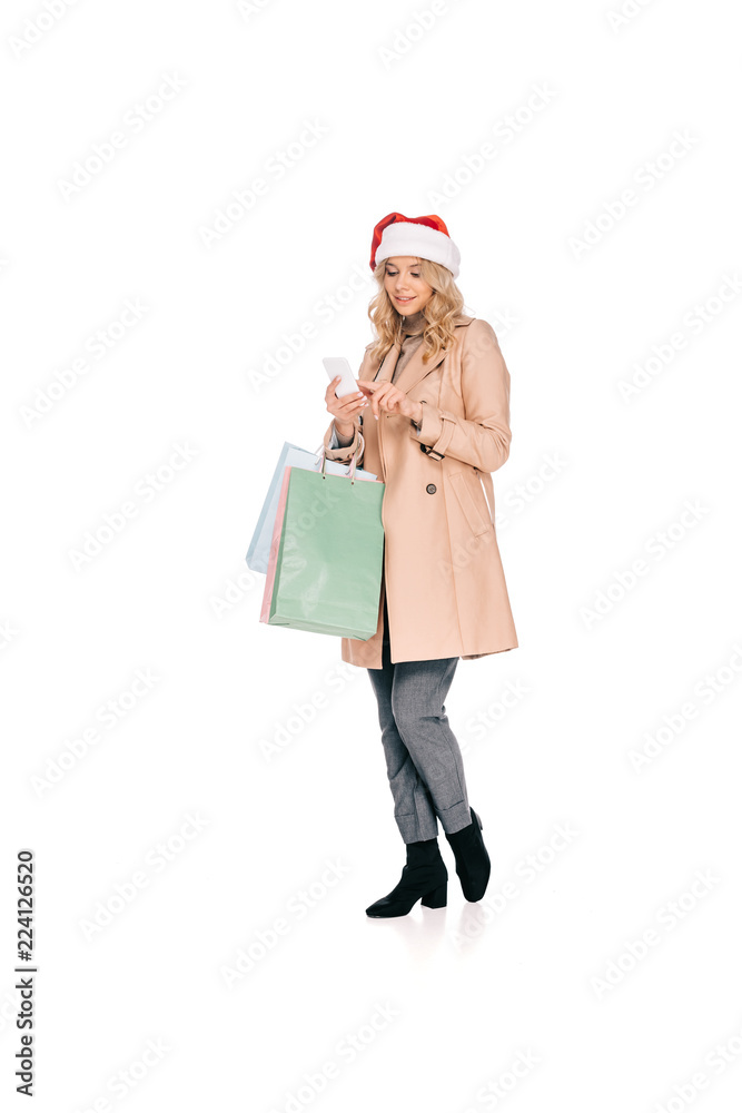 smiling young woman in santa hat holding shopping bags and using smartphone isolated on white
