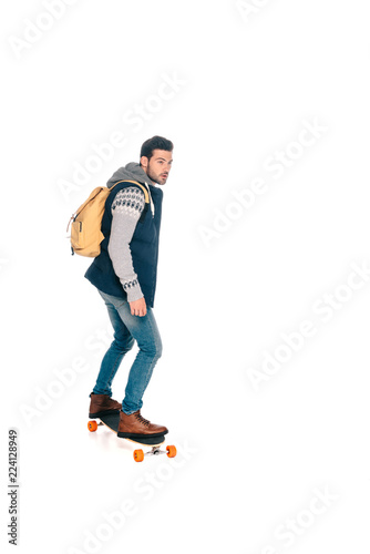 full length view of handsome young man with backpack riding longboard and looking away isolated on white