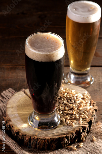 A glass of dark and light beer on a wooden stand with barley grains on a brown wooden table