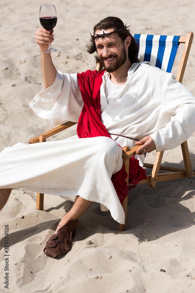 cheerful Jesus in robe and red sash resting on sun lounger and showing  glass of red wine in desert Photos | Adobe Stock