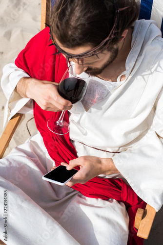 high angle view of Jesus resting on sun lounger  drinking wine and using smartphone with blank screen in desert