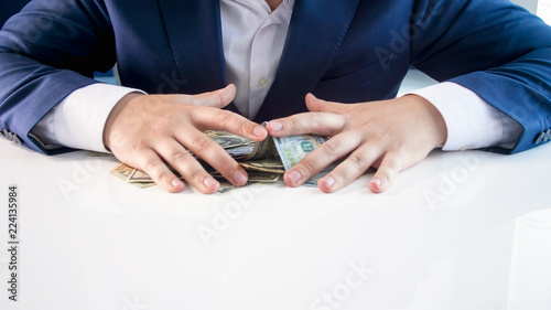 Closeup image of greedy businessman grabbing and holding lots of money on office desk