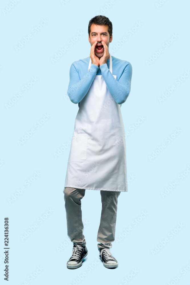 Full body of Man wearing an apron shouting with mouth wide open and announcing something on blue background Full body of Man wearing an apron shouting with mouth wide open and announcing something on blue background