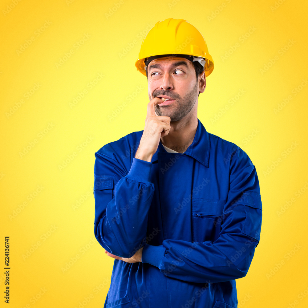 Young workman with helmet having doubts and with confuse face expression while looking up. Questioning an idea on yellow background Young workman with helmet having doubts and with confuse face expression while looking up. Questioning an idea on yellow background