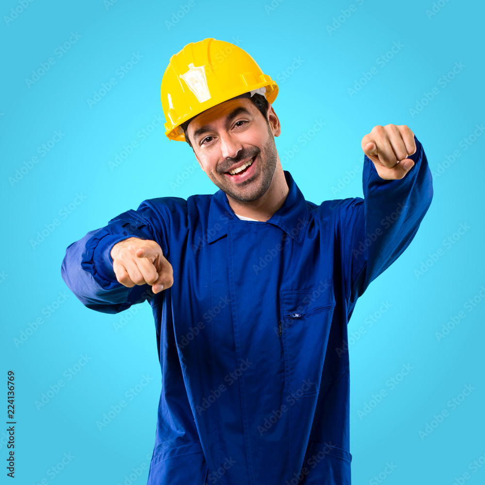 Young workman with helmet points finger at you while smiling on blue background