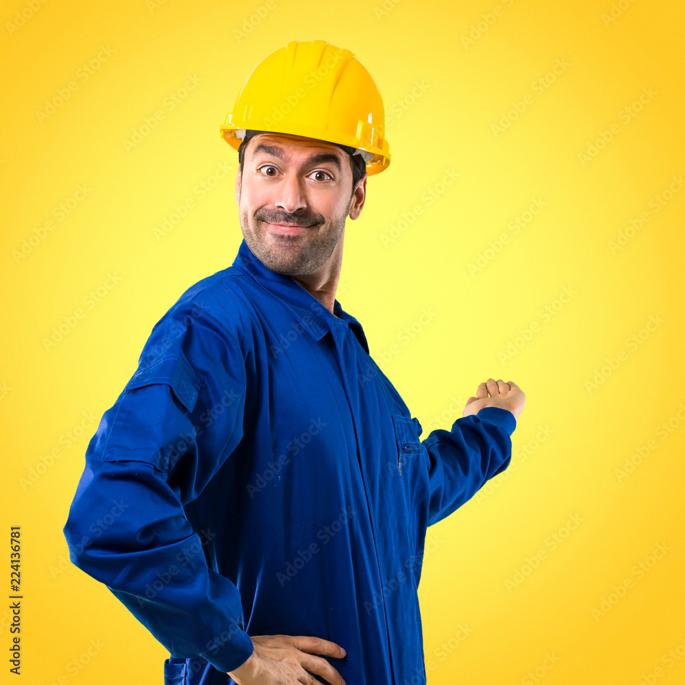 Young workman with helmet pointing back with the index finger presenting a product on yellow background Young workman with helmet pointing back with the index finger presenting a product on yellow background