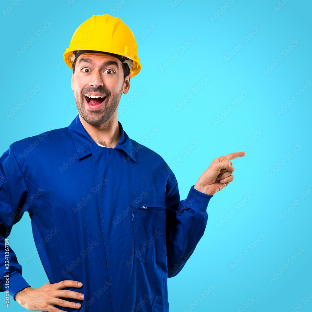 Young workman with helmet pointing finger to the side and presenting a product while smiling in a confident pose on blue background