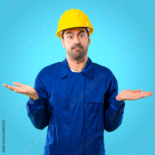 Young workman with helmet having doubts and with confuse face expression while raising hands and shoulders Uncertain concept on blue background photo