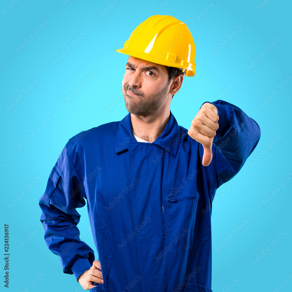 Young workman with helmet showing thumb down sign with negative expression on blue background