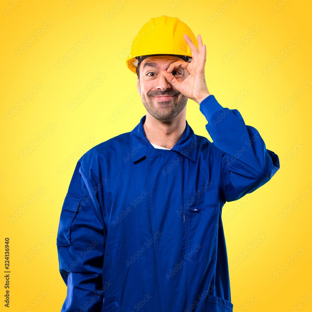 Young workman with helmet makes funny and crazy face emotion on yellow background