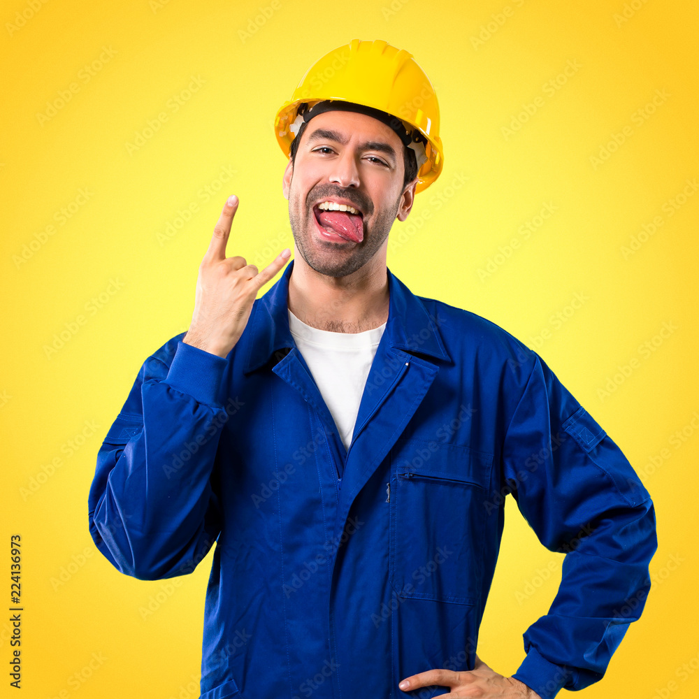 Young workman with helmet showing tongue at the camera having funny look and taking out the horns on yellow background Young workman with helmet showing tongue at the camera having funny look and taking out the horns on yellow background