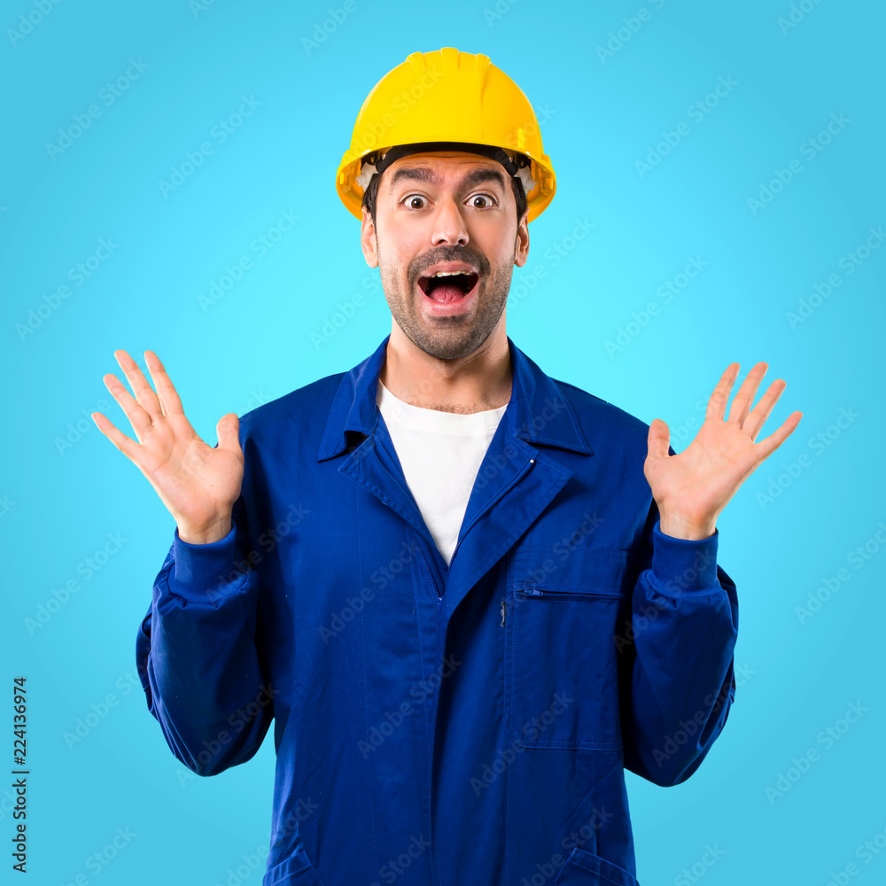 Young workman with helmet with surprise and shocked facial expression. Mouth open because not expect what has happened on blue background Young workman with helmet with surprise and shocked facial expression. Mouth open because not expect what has happened on blue background