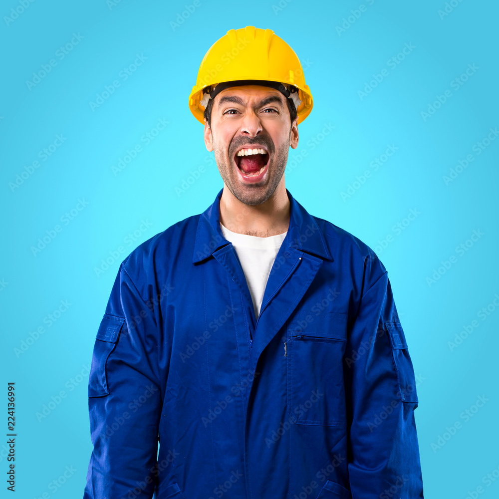 Young workman with helmet shouting to the front with mouth wide open on blue background