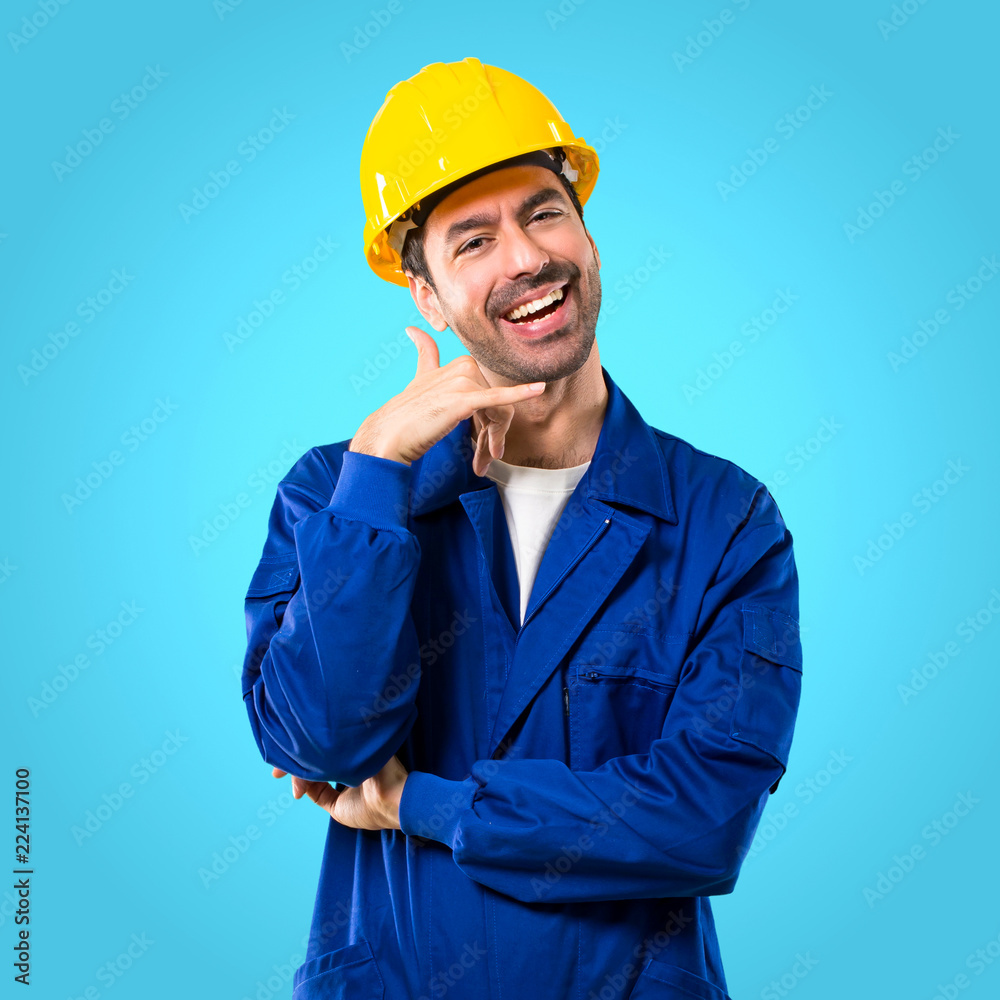 Young workman with helmet making phone gesture and speaking with someone. Call me back sign on blue background Young workman with helmet making phone gesture and speaking with someone. Call me back sign on blue background