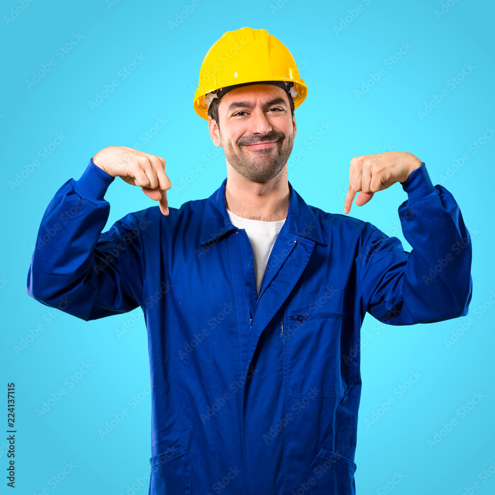 Young workman with helmet pointing down with fingers on blue background Young workman with helmet pointing down with fingers on blue background